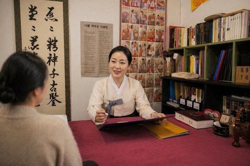 A person in traditional attire sitting at a table with books and a poster in the background. 2