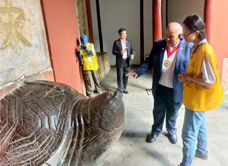 A large elephant statue with visible wear and tear, a person in a suit pointing at it, and another person in a yellow vest standing nearby. 4