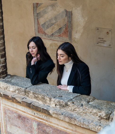 Two women standing by a stone wall with a historical plaque and a wall mural. 2