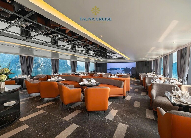 A modern cruise ship dining area with orange and grey seating, a large screen displaying a scenic view, and the 'TALIIA CRUISE' logo on the ceiling. 3
