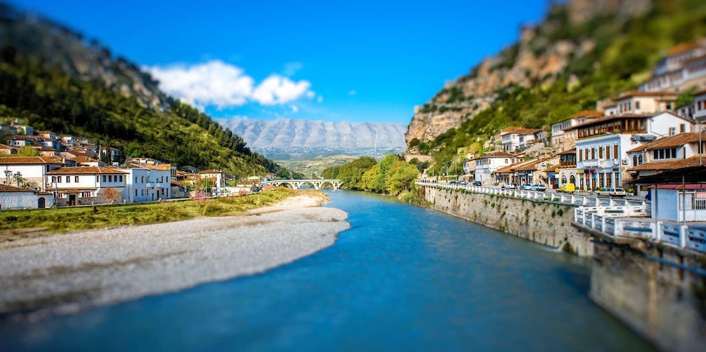A river flowing through a town with buildings on both sides, a bridge in the distance, and mountains in the background. 1