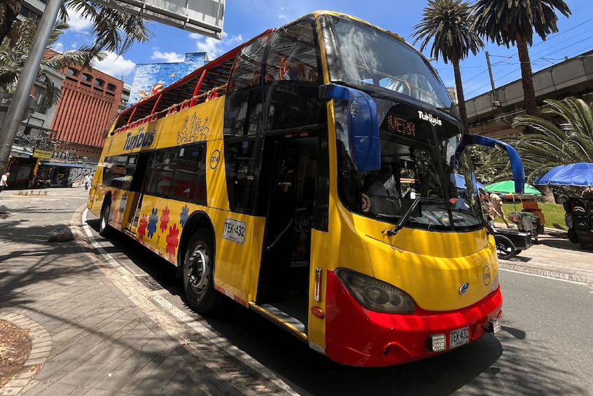 A yellow and red double-decker bus with 'Tumbis' written on its side, parked on a city street with palm trees and buildings in the background. 2