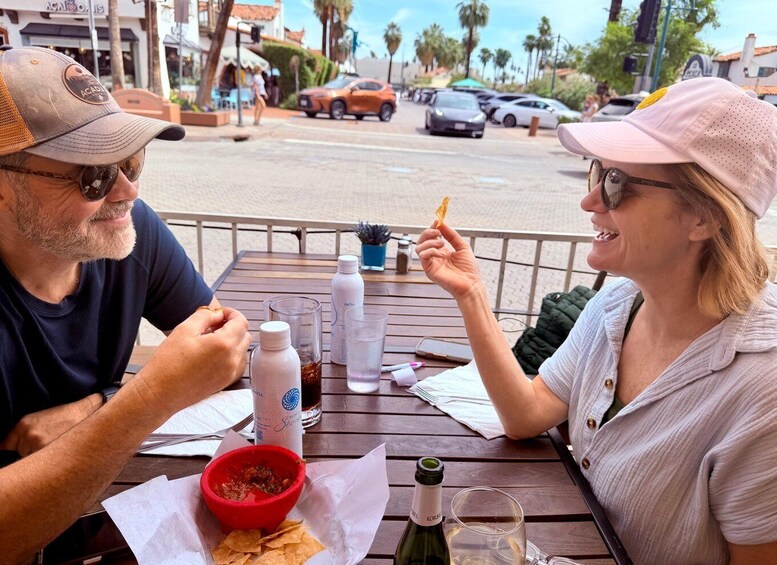 Two people dining at an outdoor table with a red bowl of food, water bottles, and a bottle of wine. 2