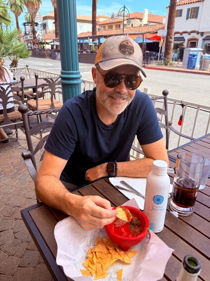 A person sitting at an outdoor table with a red bowl of food, a bottle, and a glass. 4
