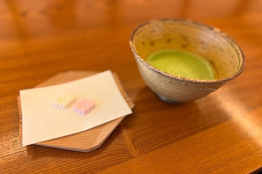 Una mesa de madera con una taza de cerámica que contiene un líquido verde y un pequeño papel doblado con un diseño blanco y rosa. 2