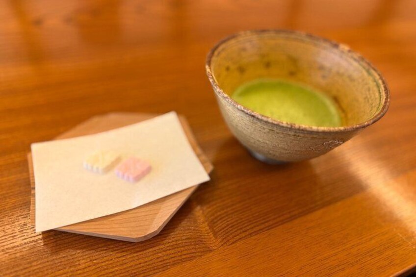 A wooden table with a ceramic cup containing a green liquid and a small folded piece of paper with a pink object on it. 4
