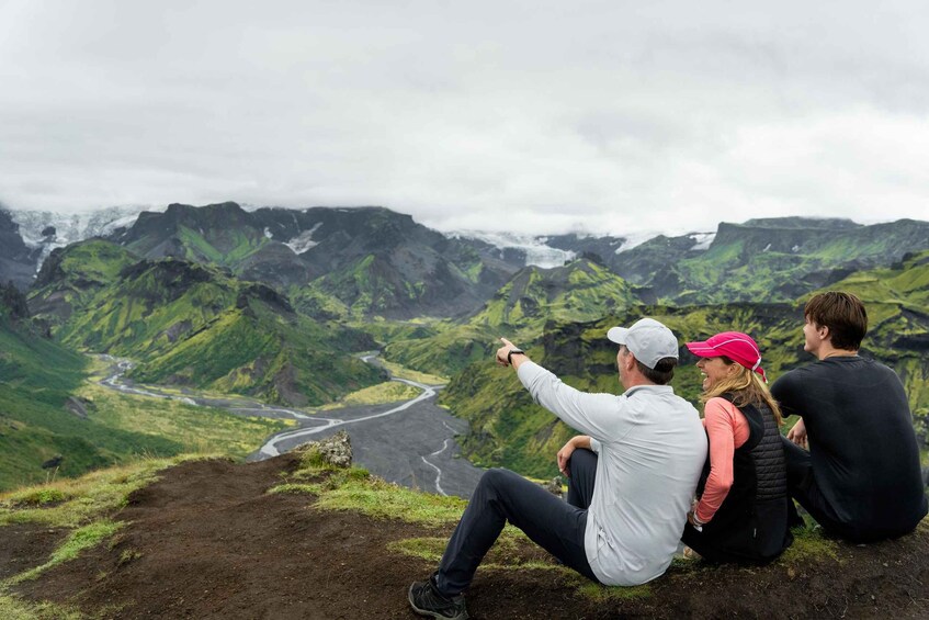 Three people sitting on a mountain ledge overlooking a river valley with green hills and a glacier in the distance. 1