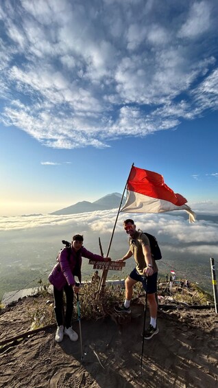 Two hikers on a mountain peak with a flag, surrounded by clouds and a clear sky. 1