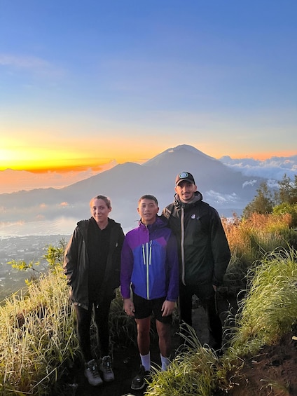 Three people standing on a grassy hill with a mountain in the background during sunset. 3
