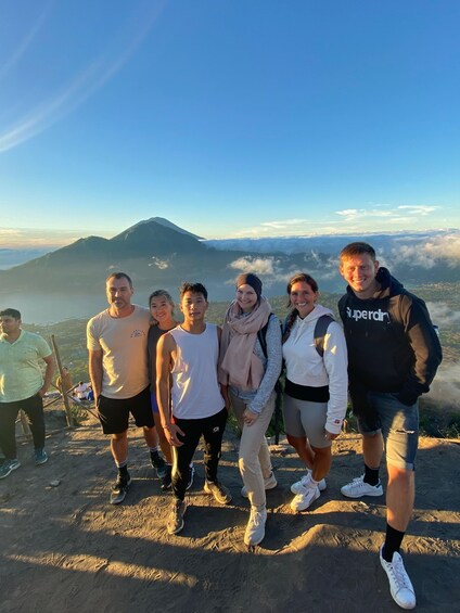A group of people standing on a mountain ridge with a view of a volcano and clouds below. 4