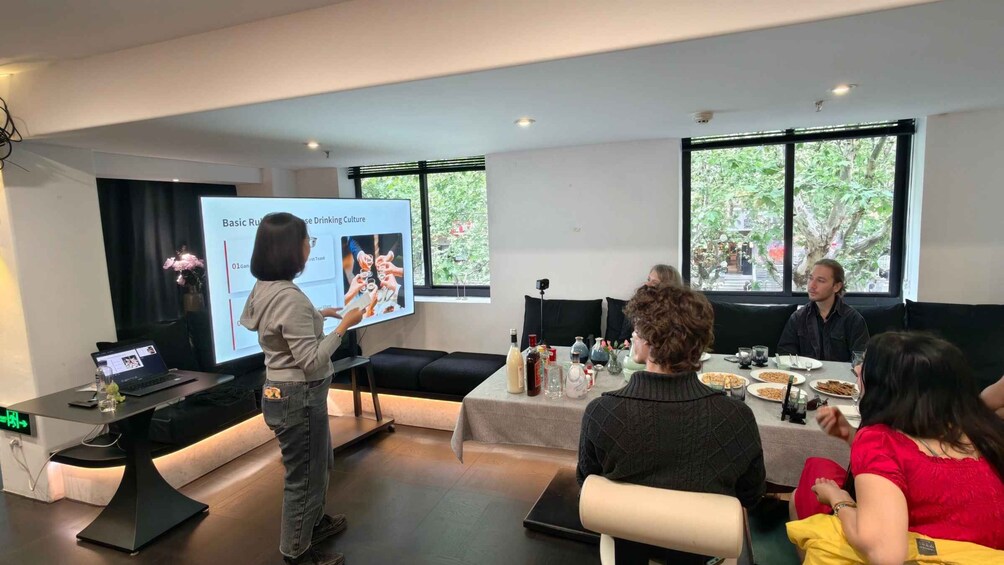 A presentation in a modern room with a projector, a laptop, and attendees seated at a table with food and drinks. 4
