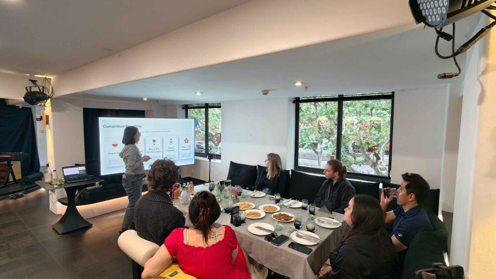 A presentation in a modern conference room with a projector screen, seated attendees, and a table with food and drinks. 5