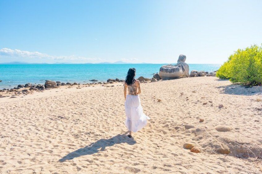 A woman in a white dress walking on a sandy beach with rocks and a green bush. 4