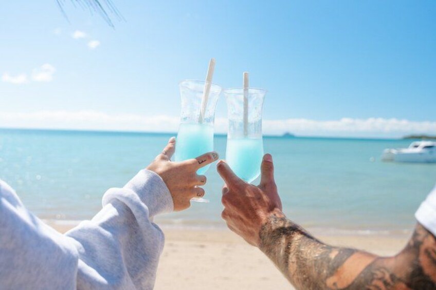 Two people toasting with blue drinks on a beach with a boat in the background. 5
