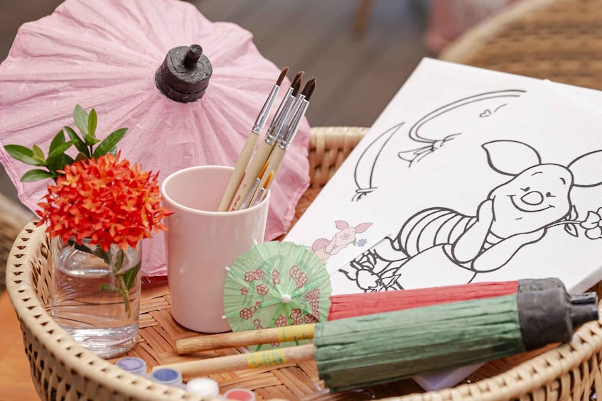 A basket with a pink umbrella, paintbrushes, a cup, a vase with flowers, and a drawing of a rabbit. 1