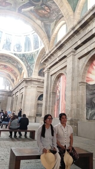 A grand architectural interior with high arches, detailed frescoes, and a group of people seated on benches. 4