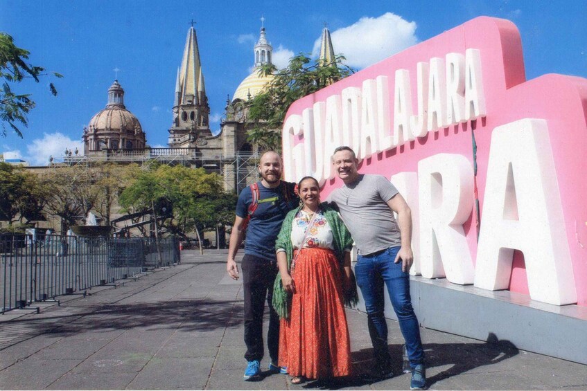 A group of people standing in front of a large pink sign with 'GUJARAS' written on it, with a historic building in the background. 2