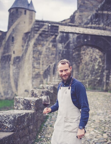 A person in a blue shirt and beige apron holding a wine glass, standing in front of a stone bridge and a castle-like structure. 2