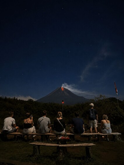Un grupo de personas sentadas en bancos de madera por la noche, observando un volcán con humo y una luz roja en la cima. 2