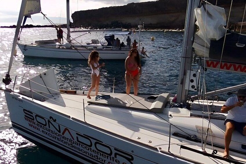 A sailboat named 'SONADOP' with people on deck, calm water, and a rocky coastline in the background. 5