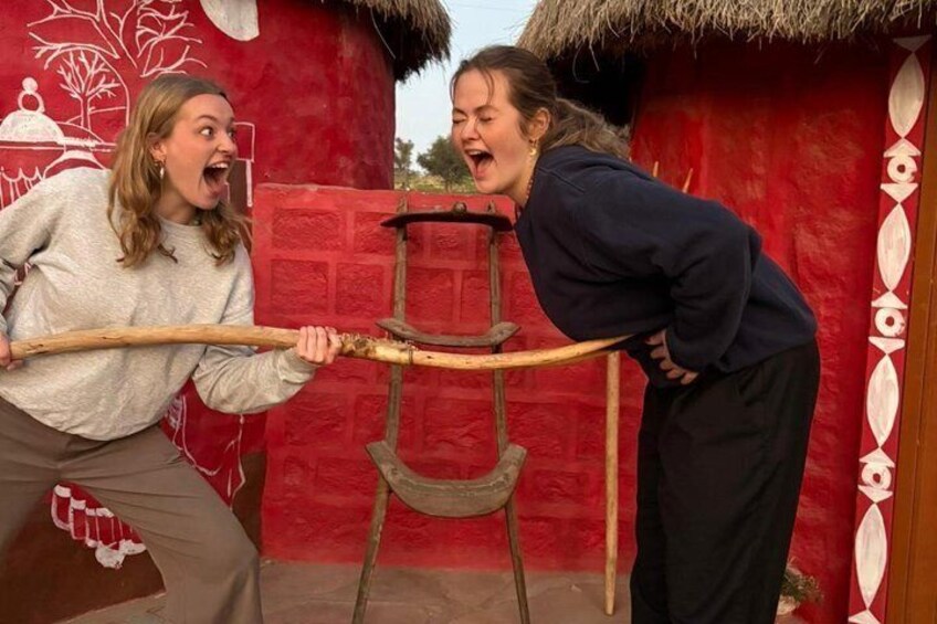 Two people playing tug-of-war with a wooden stick in front of a red building with a thatched roof. 4