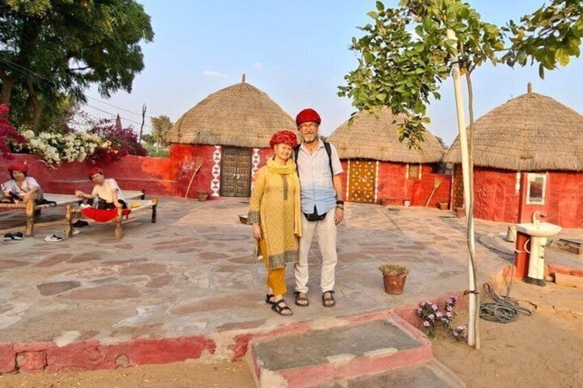 Two people standing in front of traditional thatched-roof huts with a stone-paved courtyard. 5