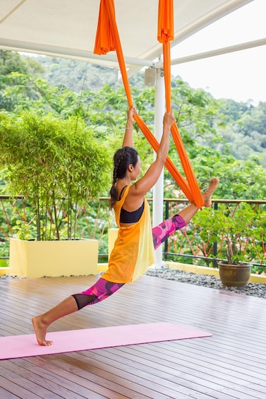 A person performing aerial yoga on a pink mat outdoors with greenery in the background. 4