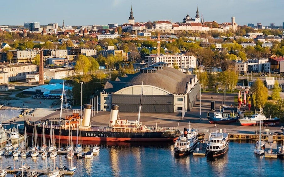 A harbor with a large ship named 'SUUR TOLL' docked, surrounded by smaller boats and a cityscape with historic buildings in the background. 3