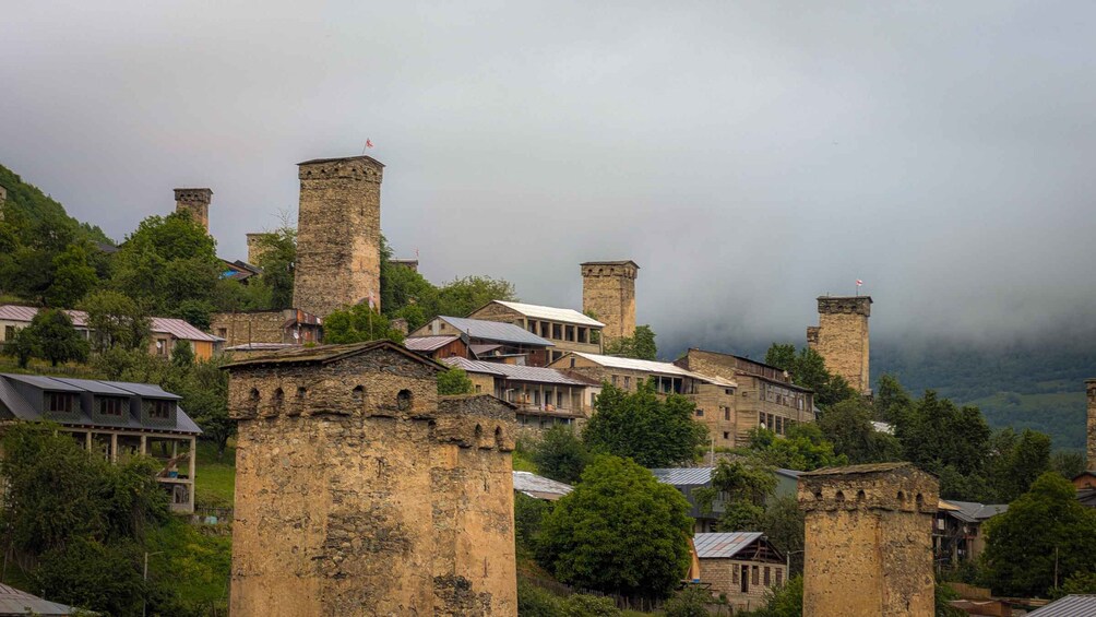 A village with stone buildings and towers, surrounded by trees and hills. 1
