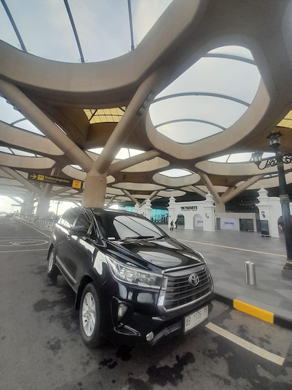 A black SUV parked at an airport terminal with a distinctive modern architectural design featuring circular skylights and a sign indicating the departure area. 1