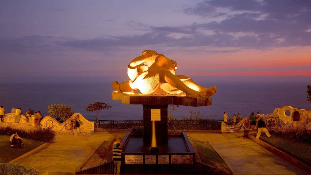 A large illuminated sculpture of a reclining figure, surrounded by a paved area with people walking around. 2