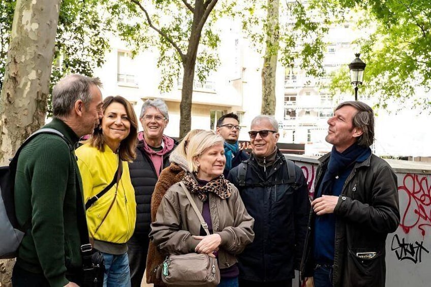 A group of people standing outdoors, some wearing jackets, with trees and a building in the background. 1
