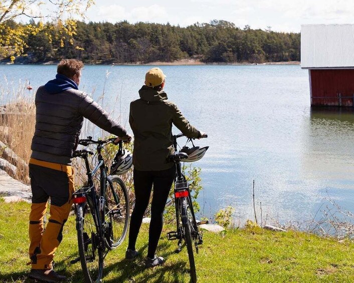 Two people standing by a lake with bicycles, a red building in the background, and a forested shoreline. 1