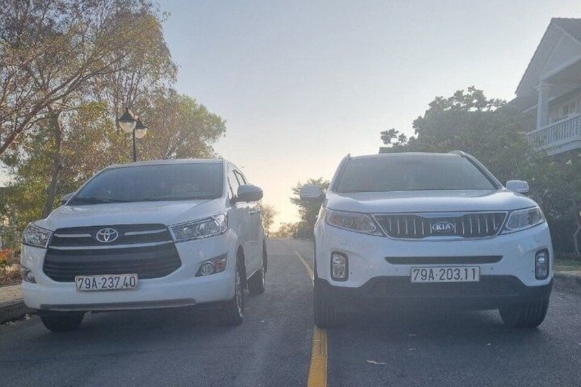 Two white SUVs parked on a street with trees and a building in the background. 1
