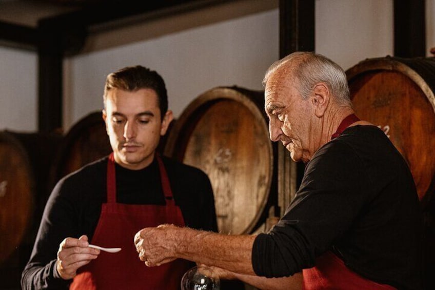 Two individuals in a cellar with wooden barrels, one holding a spoon. 1
