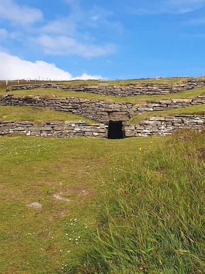 A stone structure with a central opening, surrounded by grassy terrain and a clear sky. 4