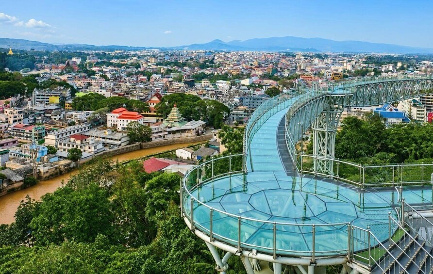A cityscape with a prominent glass bridge over a river, surrounded by dense greenery and urban buildings. 1