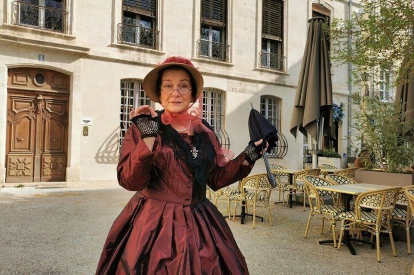 A person in a red dress and hat standing in front of a building with a large wooden door and windows. 1