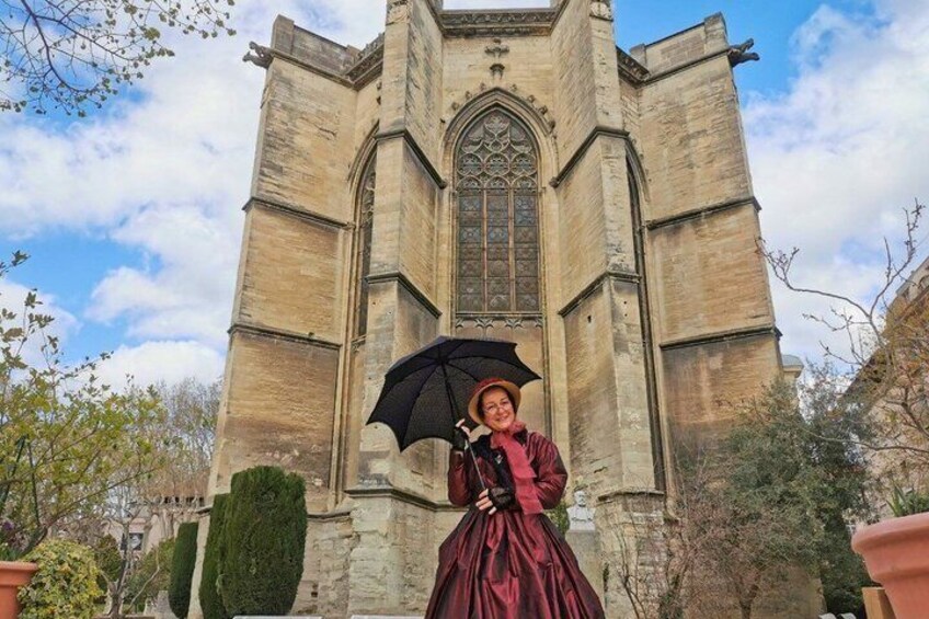 A person in a red dress standing in front of a historic stone building with a large arched window and a pointed roof. 2