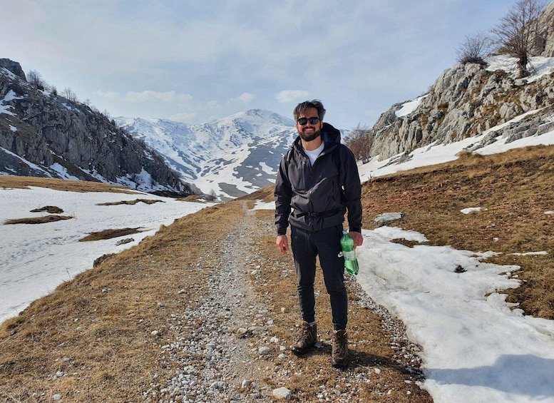 A person standing on a gravel path with snow patches and rocky mountains in the background. 5