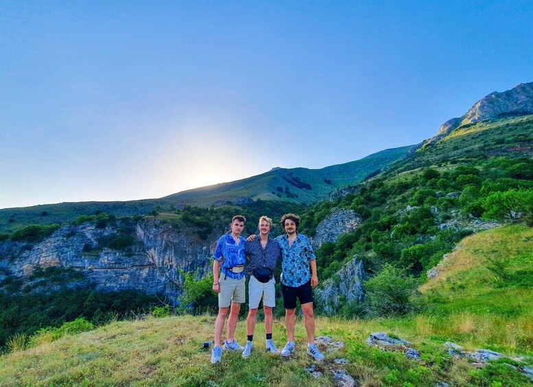 Three people standing on a grassy hill with a mountainous landscape in the background. 2