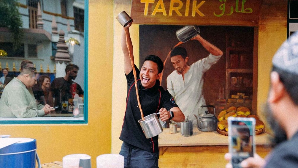 A street food vendor pouring drinks, a customer taking a photo, and a shop with a sign reading 'TARIK'. 1