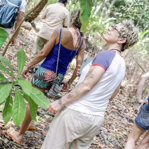 A person with tattoos in a forested area, wearing a white shirt and shorts, with another person in the background. 2