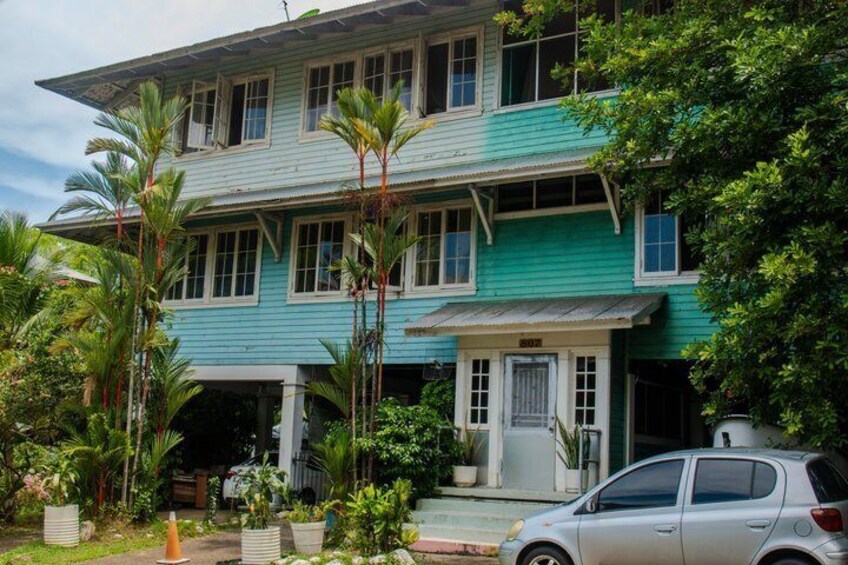 A two-story house with teal siding, a car parked in front, and tropical vegetation. 3