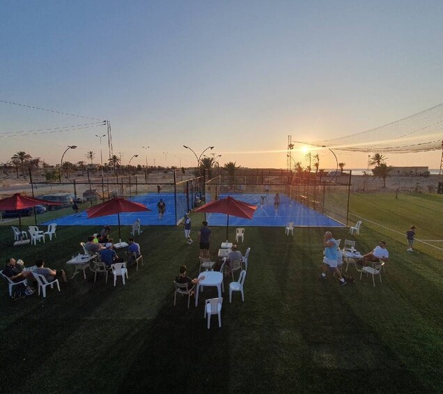 An outdoor sports court with people playing, surrounded by chairs and umbrellas. 1