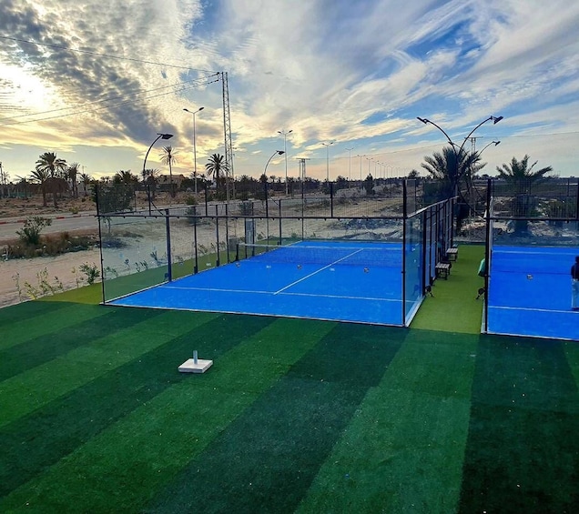 A tennis court with a blue surface and green artificial turf, surrounded by a fence and palm trees. 2