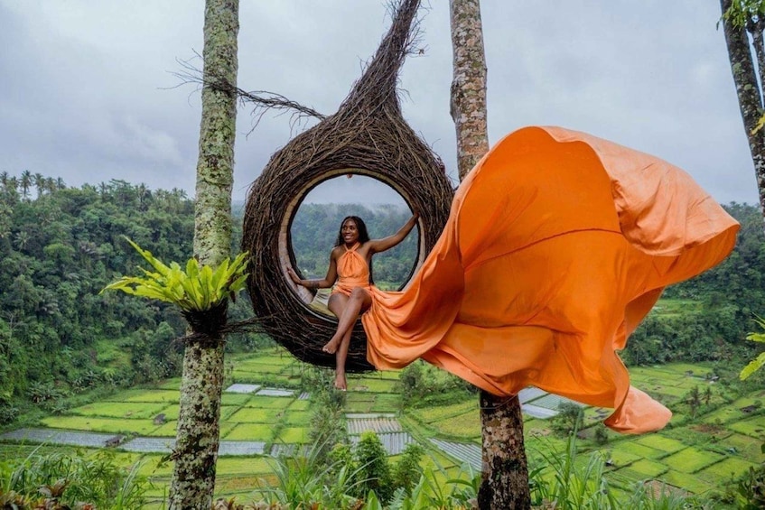 A person in an orange dress sitting in a circular wicker structure amidst a lush green landscape with terraced fields. 4
