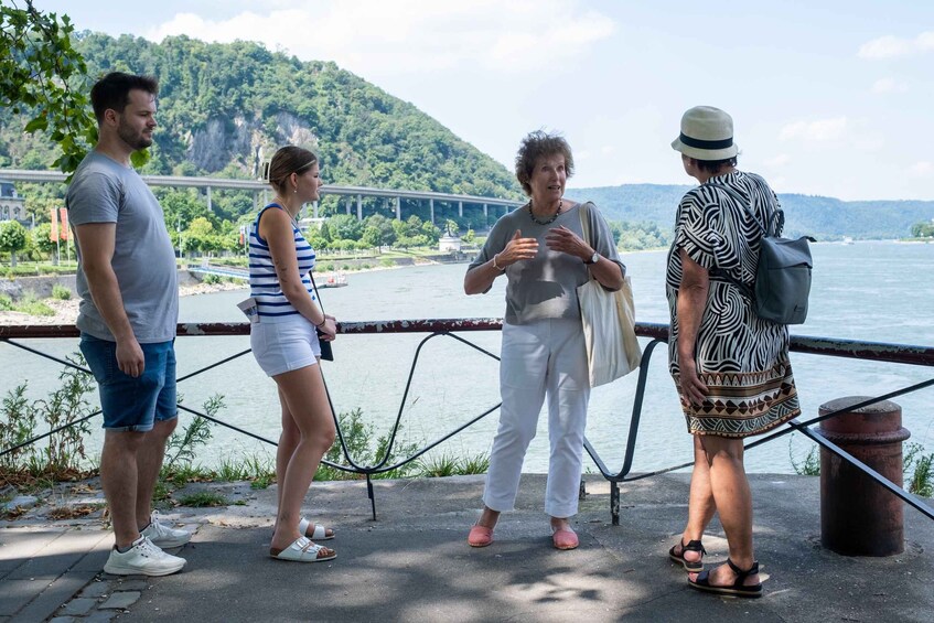 A group of people standing by a riverside walkway with a bridge and hills in the background. 2