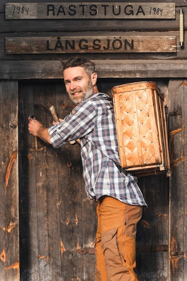 A person with a wooden case on their back standing in front of a wooden door with signs reading '1984 RASTSTUGA LÄNGSJÖN'. 2