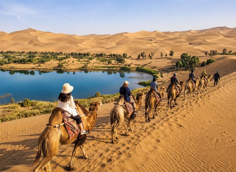 A group of people riding camels along a sandy path beside a lake in a desert landscape. 4